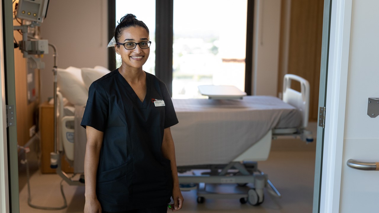 Radhika John stands smiling in front of a patient room in the Victorian Heart Hospital. She wears navy scrubs, black glasses and wears her black hair in a top bun.