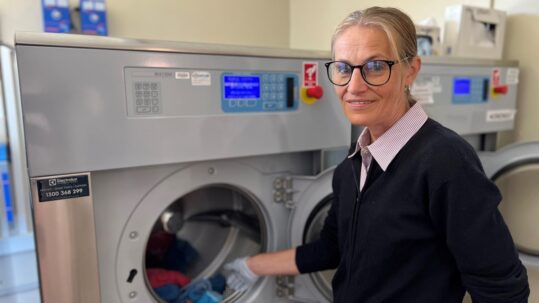 Valerie Villinger stands in front of an industrial washing machine. She is placing some washing inside the drum while smiling at the camera.